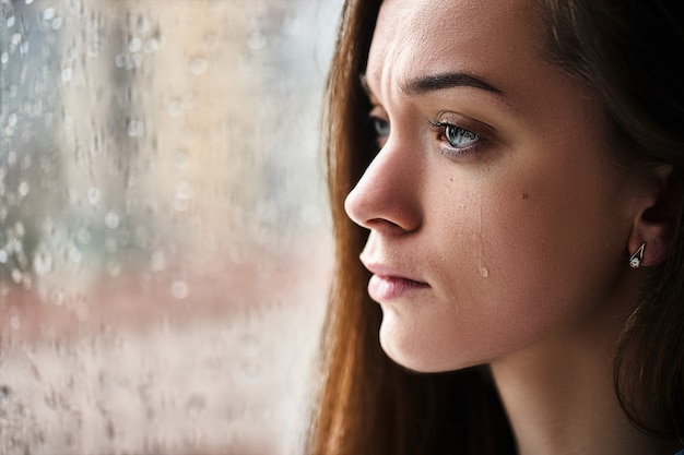 Photo sad upset crying woman with tears eyes suffering from emotional shock, loss, grief, life problems and break up relationship near window with raindrops. female received bad news