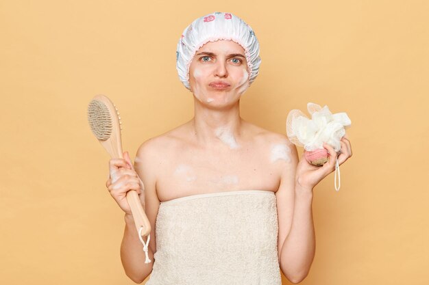 Photo sad upset disappointed woman wearing shower cap standing isolated over beige background holding washcloth and brush for dry massage