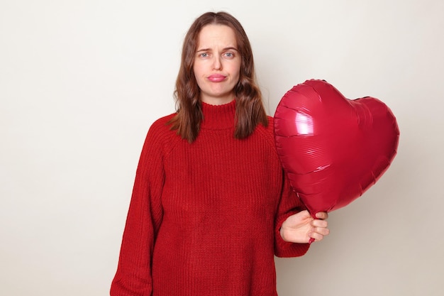 Sad upset woman with brown hair wearing red jumper standing isolated over gray background with heart shaped balloon has not mood to celebrate Valentines Day expressing sorrow and sadness