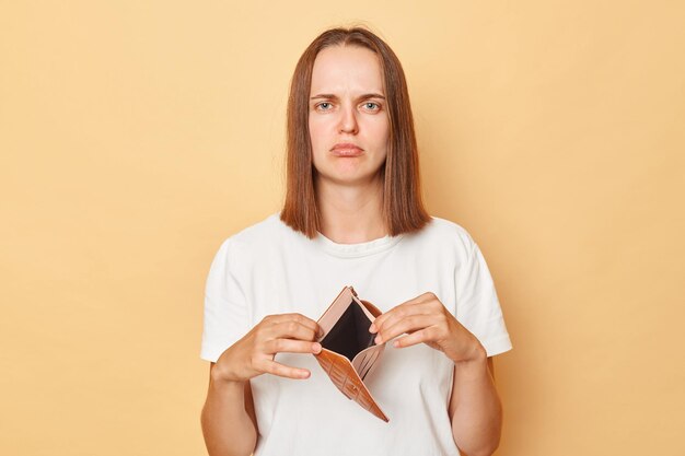 Sad upset young caucasian woman wearing white Tshirt showing empty wallet over isolated beige background has no money to spend looking at camera with displeased expression