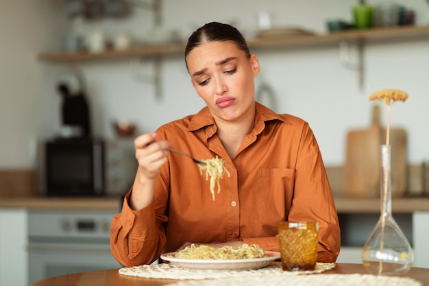 Sad upset young european woman sitting at table in kitchen interior looking at delivered pasta and