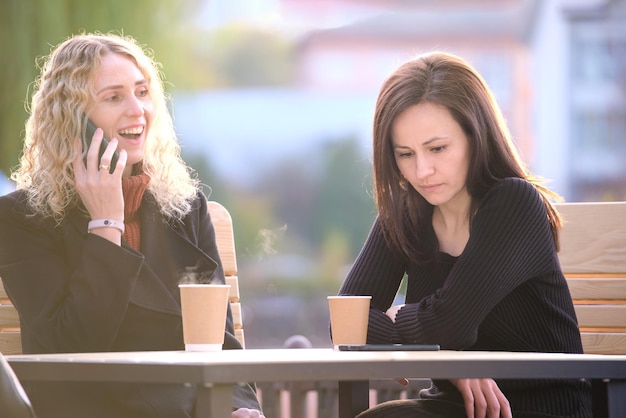 Sad woman being ignored by her friend sitting at street cafe outdoors while she is talking happily on mobile phone and paying no attention Friendship problems concept