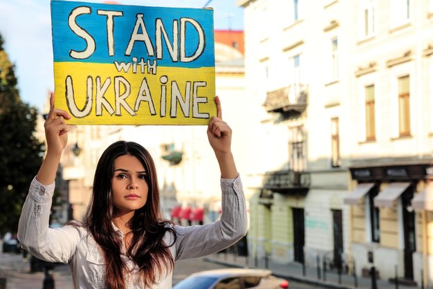 Photo sad woman holding poster in colors of national flag and words stand with ukraine on city street space for text