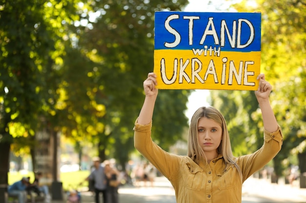 Photo sad woman holding poster in colors of national flag and words stand with ukraine outdoors space for text