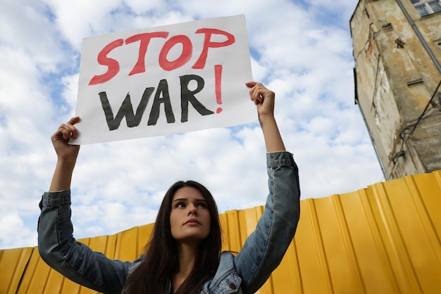 Sad woman holding poster Stop War near yellow fence under blue cloudy sky