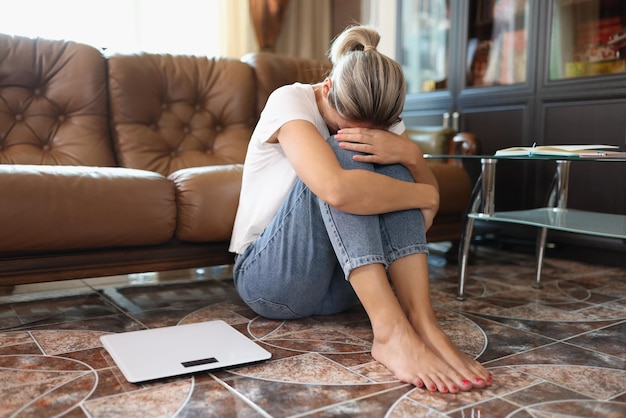 Sad woman sits on floor near scales and holding her knees by hands