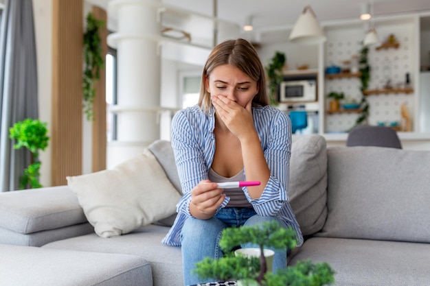 Sad worried woman checking her recent pregnancy test sitting on couch at home Maternity child birth and family problems concept unwanted pregnancy