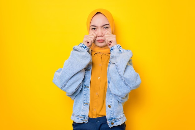 Sad young Asian woman in jeans jacket rubs tears wants to cry depressed has a problem isolated over yellow background