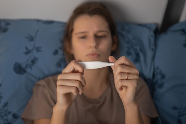 Sad young caucasian girl lying on pillow and looking on thermometer. Sickness woman hold temperature meter in hands. Female suffering from disease, virus or infection. Selective focus. Healthcare.