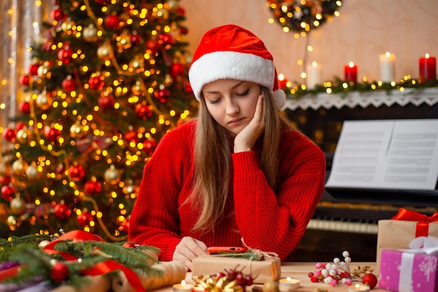 Sad young girl in red sweater and santa hat sits at the table and watches something on smartphone Festive decoration of the room but bad mood of person