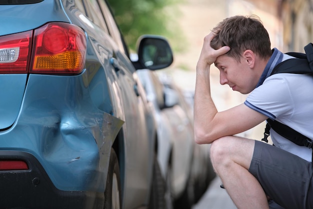 Sad young man driver sitting near his dented car looking shocked on damaged fender in road accident