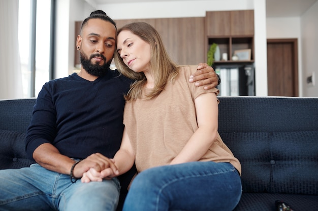 Sad young mixed-race couple reassuring each other when sitting on sofa at home