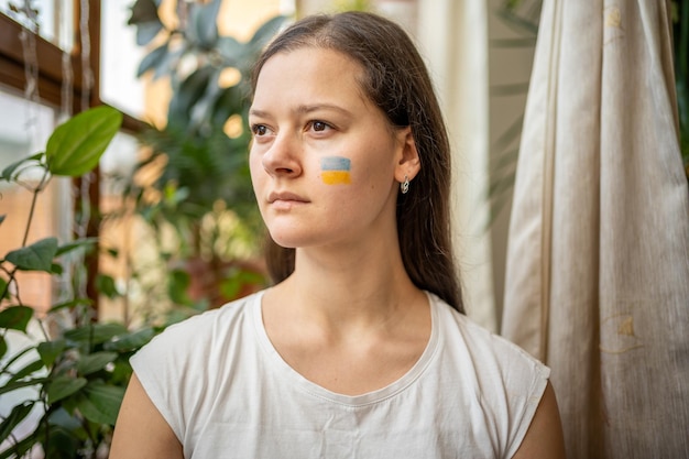 A sad young ukrainian girl with the flag of ukraine on her face is looking through window the concep