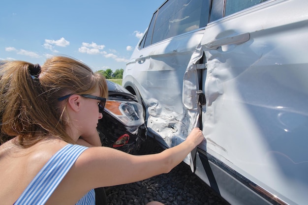 Sad young woman driver sitting near her smashed car looking shocked on crashed vehicles in road accident