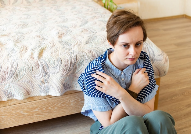 A sad young woman folded her arms across her chest. Sits on the floor in his bedroom at home