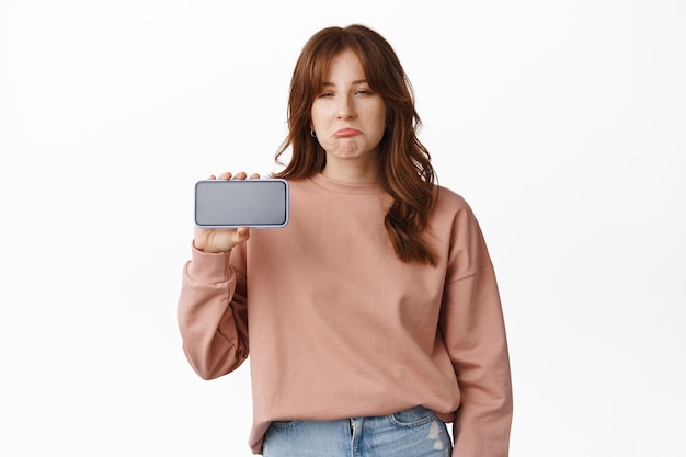 Sad young woman sobbing, look and show smartphone screen, staring at unfair thing, disappointed by mobile phone app, standing on white