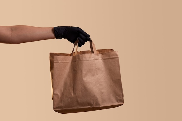 Safe delivery for quarantine female hand in black latex glove gives paper bag over beige wall