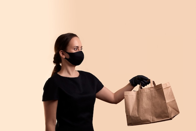 Safe delivery for quarantine woman in black dress face mask and latex gloves gives paper bag over beige wall