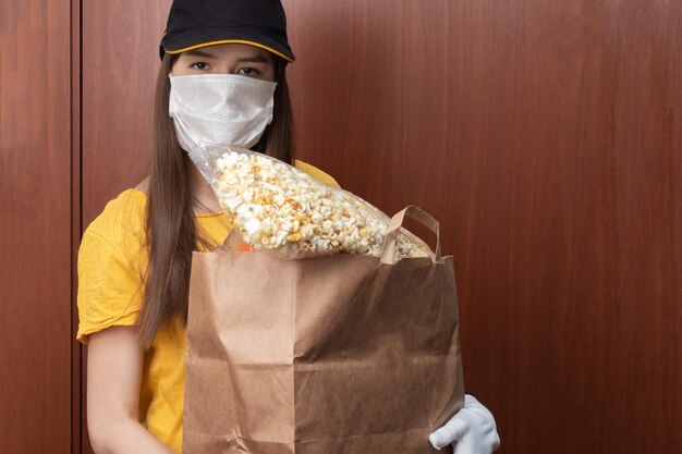 Safe Food Delivery and Online Orders Young woman courier in uniform in a medical antibacterial mask and gloves stands at the door with a package