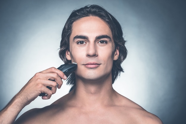 Safe shaving. Handsome young shirtless man shaving with electric razor and looking at camera while standing against grey background