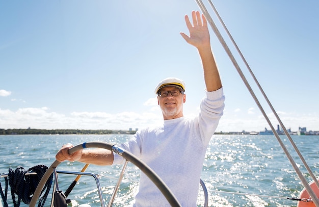 sailing, age, tourism, travel and people concept - happy senior man in captain hat on steering wheel and waving hand sail boat or yacht floating in sea