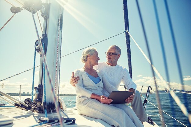 Photo sailing, technology, tourism, travel and people concept - happy senior couple with tablet pc computer talking on sail boat or yacht deck floating in sea