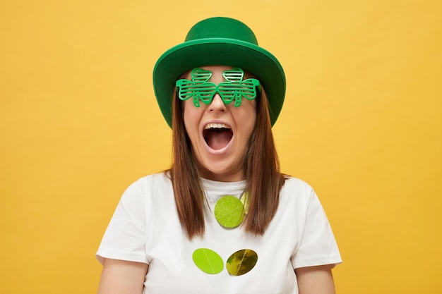 Saint Patricks Day party Extremely happy overjoyed woman wearing green hat and clover glasses standing isolated over yellow background screaming with excitement rejoicing