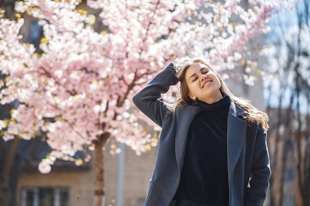 Sakura branches with flowers on a tree on the city streets. Happy woman girl in a gray palette walks along an alley with blooming sakura. Gorgeous fancy girl outdoors. Sakura tree blooming.