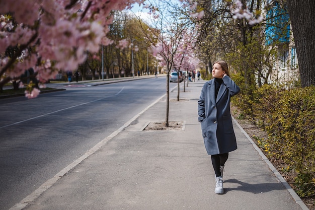 Sakura branches with flowers on a tree on the city streets. Happy woman girl in a gray palette walks along an alley with blooming sakura. Gorgeous fancy girl outdoors. Sakura tree blooming.