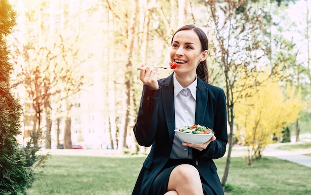 Salad on business lunch Portrait of a beautiful modern and smiling business woman in a black suit with a plate of vegetable salad in the hands that she eats on the street near work