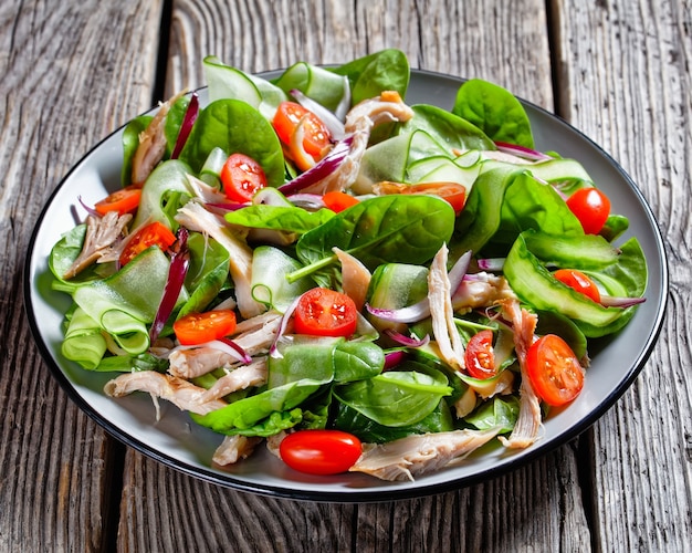 Salad of chicken breast and fresh baby spinach leaves, cherry tomatoes, cucumber ribbon, red onion rings with olive oil and lemon dressing, served on a plate on a wooden table, top view, close-up