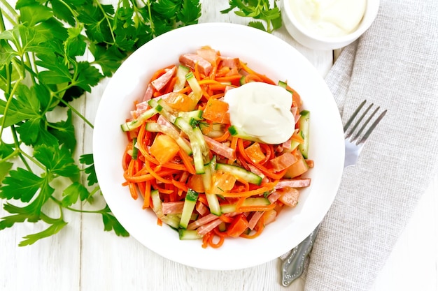 Salad from smoked sausage spicy carrot tomato cucumber and spices with mayonnaise napkin fork and parsley on a light wooden board background from above