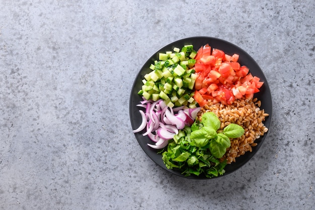 Salad of whole grain cereal spelt with vegetables, tomato, cucumber, greens on grey stone table with 