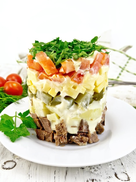 Salad with beef boiled potatoes pickles cheese tomato parsley in a white plate napkin background dill on a wooden board