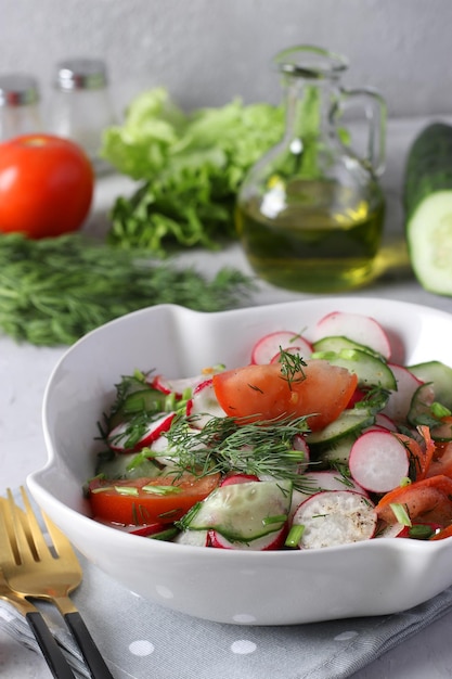 Salad with cucumbers tomatoes and radishes dressed with olive oil in white salad bowl on a gray background