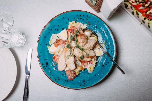 Salad with different meat tomatoes and eggs all sprinkled with parmesan on a blue plate which stands on a white tablecloth