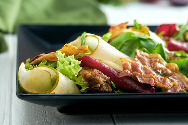 Salad with liver pear and bacon in a black plate on a wooden background