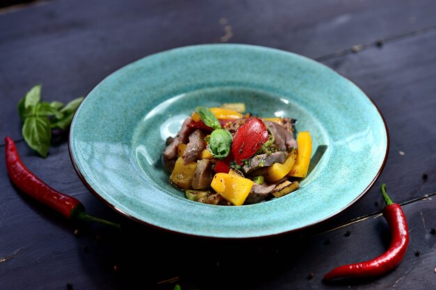 Salad with meat, bell peppers, tomatoes and basil, in a turquoise plate on a wooden stove