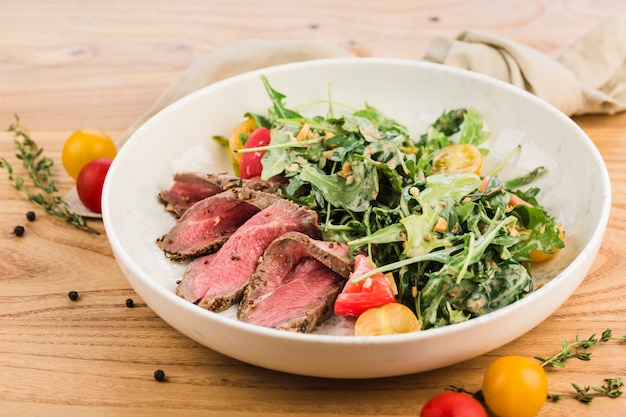 Salad with roast beef, arugula and cherry tomatoes in a plate on a light wooden surface.