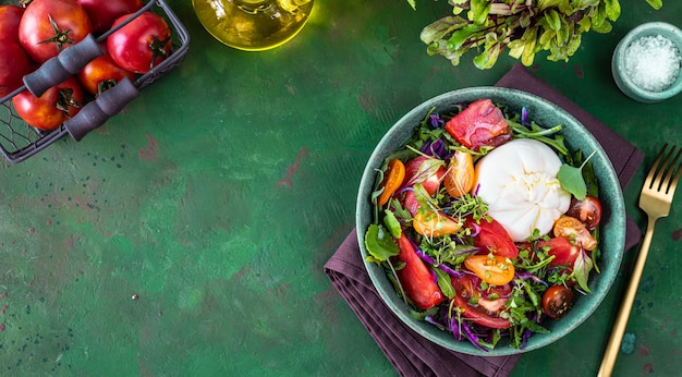 Salad with tomatoes, arugula, Burrata cheese and microgreens on a green stone background, top view. copy space