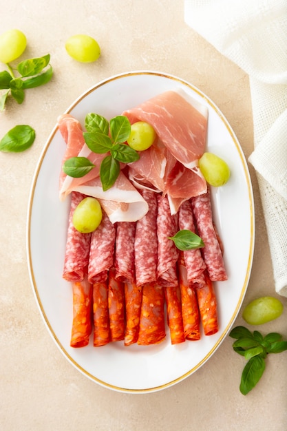 Salami and prosciutto slices in plate, with basil and grapes. Delicious snack for wine. Top view.