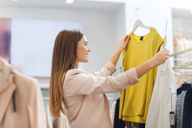 sale, clothes , shopping, fashion and people concept - happy young woman choosing between two shirts in mall or clothing store