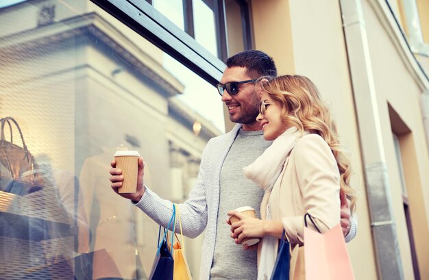 Photo sale, consumerism and people concept - happy couple with shopping bags and coffee paper cups looking at shop window in city