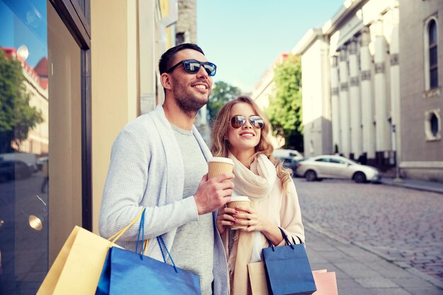 Photo sale, consumerism and people concept - happy couple with shopping bags and coffee paper cups at shop window in city