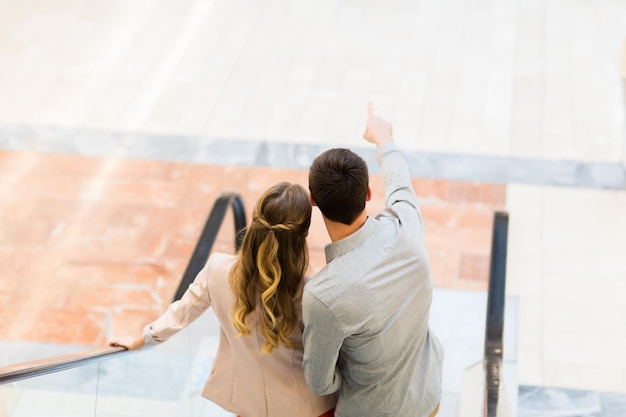 sale, consumerism and people concept - happy young couple with shopping bags going down by escalator and pointing finger in mall