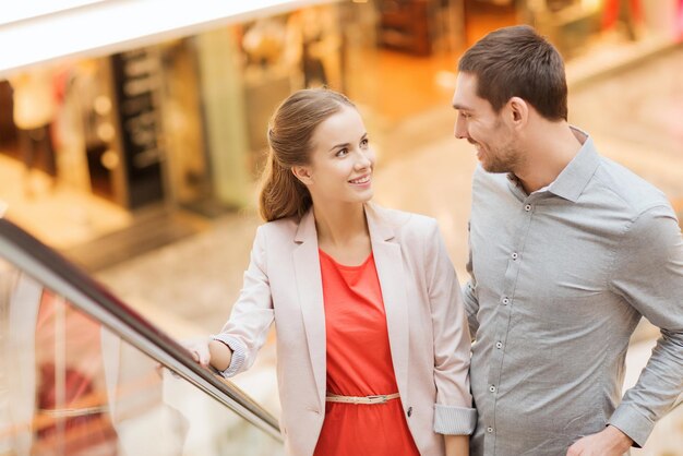 sale, consumerism and people concept - happy young couple with shopping bags rising on escalator and pointing finger in mall