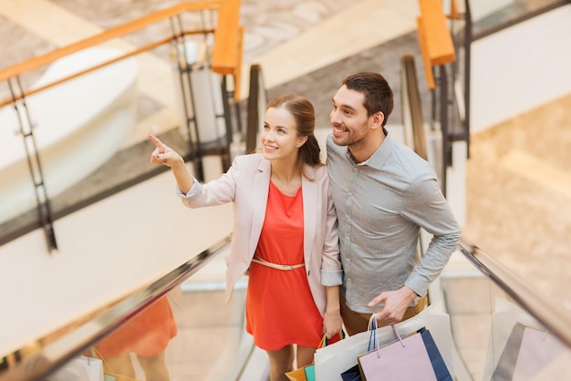 sale, consumerism and people concept - happy young couple with shopping bags rising on escalator and pointing finger in mall