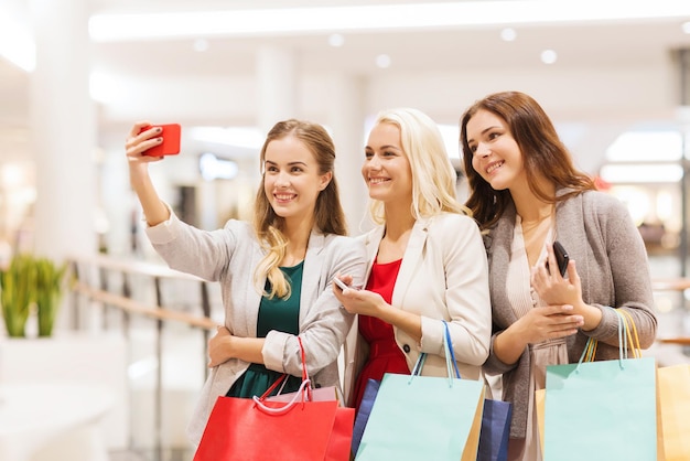 sale, consumerism, technology and people concept - happy young women with smartphones and shopping bags taking selfie in mall