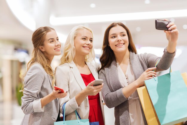 Photo sale, consumerism, technology and people concept - happy young women with smartphones and shopping bags taking selfie in mall