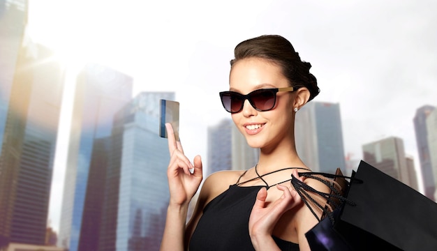 sale, finances, fashion, people and luxury concept - happy beautiful young woman in black sunglasses with credit card and shopping bags over city skyscrapers background